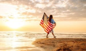 Woman Holds US Flag Walking On Beach Sunset