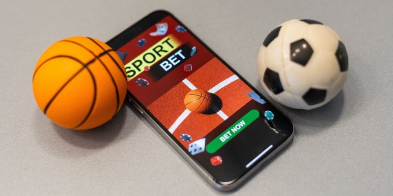 a mobile phone with sports betting on it next to a mini basketball and mini soccer ball