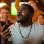 Enthused Black Man Watching Holding Mobile Phone with DraftKings Logo