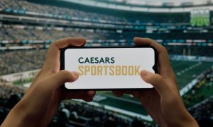 Hands Hold Cell Phone with Caesars Sportsbook Logo inside Football Stadium