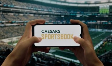 Hands Hold Cell Phone with Caesars Sportsbook Logo inside Football Stadium