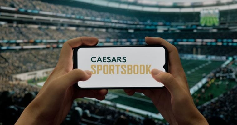 Hands Hold Cell Phone with Caesars Sportsbook Logo inside Football Stadium
