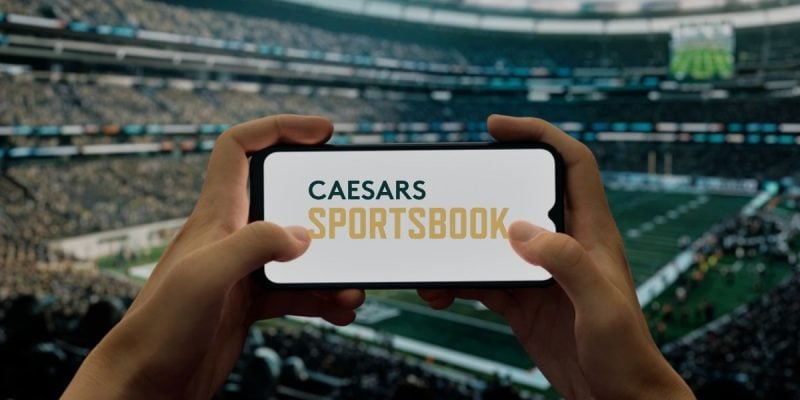Hands Hold Cell Phone with Caesars Sportsbook Logo inside Football Stadium