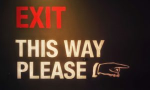 Red and White Exit Sign Reads "Exit This Way Please" With Hand Pointing