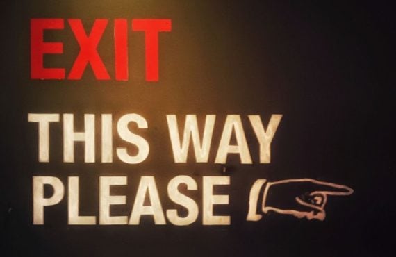 Red and White Exit Sign Reads "Exit This Way Please" With Hand Pointing