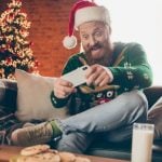 Man Sits at Home Playing on Mobile Phone with Christmas Tree Background