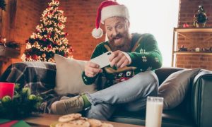 Man Sits at Home Playing on Mobile Phone with Christmas Tree Background