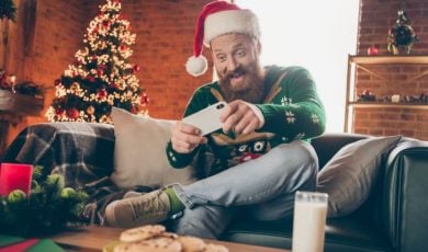 Man Sits at Home Playing on Mobile Phone with Christmas Tree Background
