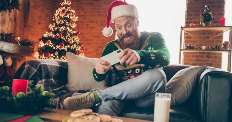 Man Sits at Home Playing on Mobile Phone with Christmas Tree Background