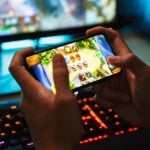 Boy Plays Online Game Using Smartphone with Desktop Computer in Background