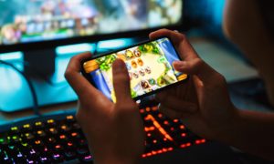 Boy Plays Online Game Using Smartphone with Desktop Computer in Background