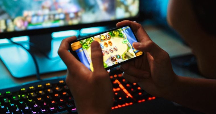 Boy Plays Online Game Using Smartphone with Desktop Computer in Background