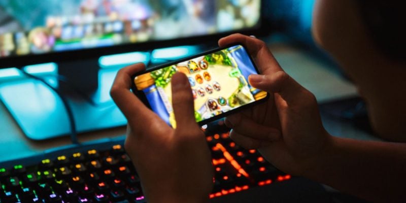 Boy Plays Online Game Using Smartphone with Desktop Computer in Background