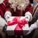 Close Up of Christmas Gift Wrapped with Red Bow Held By Three Santas