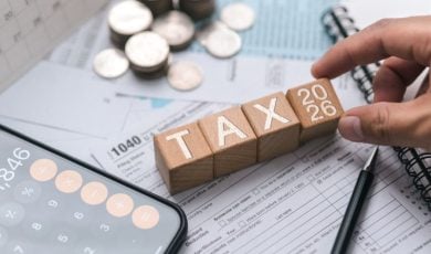 Man's Hand Touches Wood Blocks Spelling Tax 2026 Next to Calculator, Coins & Tax Forms
