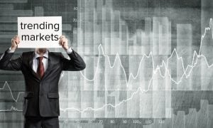 Man in Black Suit Holds Paper Reading Trending Markets In Front Of Face And Stands In Front Of Gray and White Line Graph