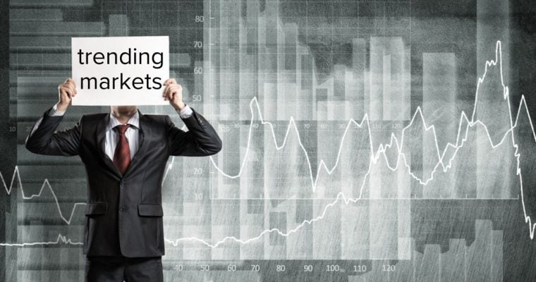 Man in Black Suit Holds Paper Reading Trending Markets In Front Of Face And Stands In Front Of Gray and White Line Graph