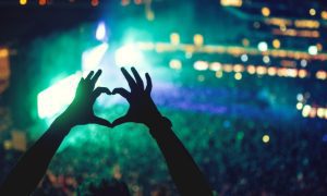 Fan Among Crowd of Fans Puts Up Hands Forming Heart While Watching Concert Displaying Bright Lights