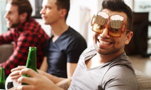 Friends Sit On Couch Watching TV, One Wears Beer Glass Shaped Glasses and Holds Green Berr Bottle