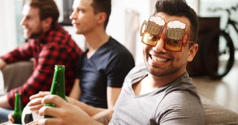 Friends Sit On Couch Watching TV, One Wears Beer Glass Shaped Glasses and Holds Green Berr Bottle