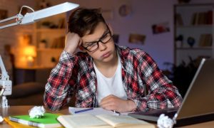 Teenage Boy Wears Glasses Sitting at Desk with Books and Laptop and Frustrated Expression