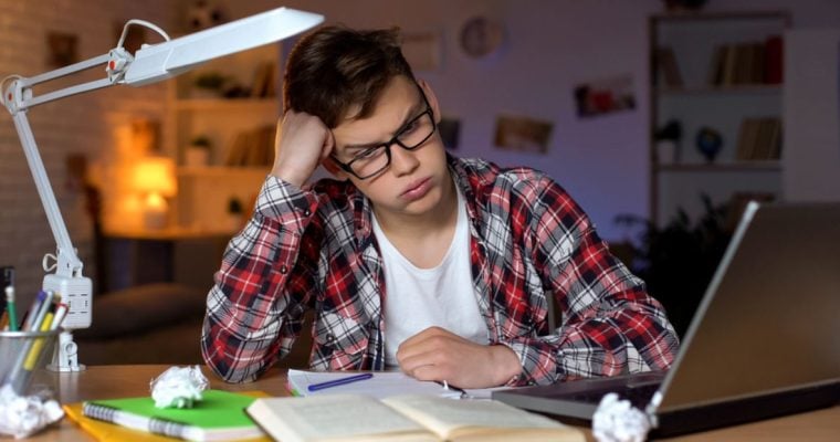 Teenage Boy Wears Glasses Sitting at Desk with Books and Laptop and Frustrated Expression