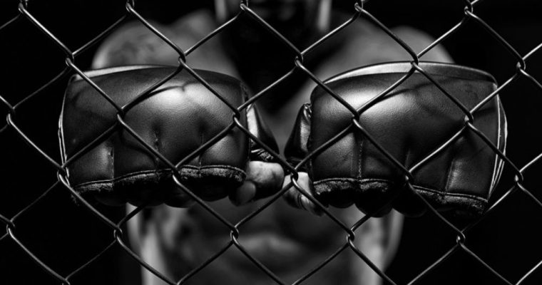 Man in MMA Fighting Cage Holds Fists with Black Gloves Up to Fence