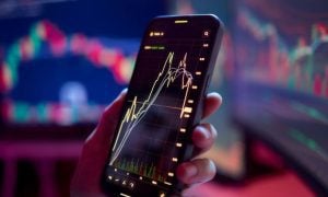 Man Holds Cell Phone With Line Graph Chart and Desktop Computers in Background