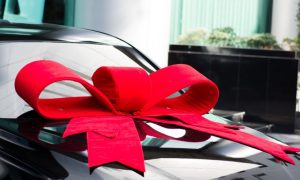 Large Red Bow Sits On Top Of Black Car in Outside Surroundings