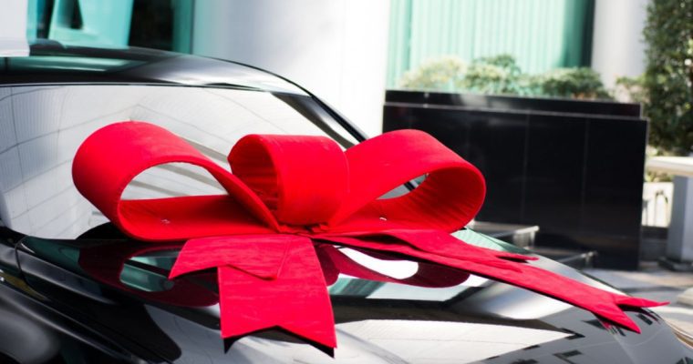 Large Red Bow Sits On Top Of Black Car in Outside Surroundings