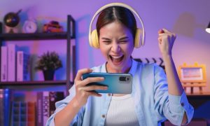 Excited Young Woman Holds iPhone Wearing Yellow Headphones Sitting at Desk Inside Room