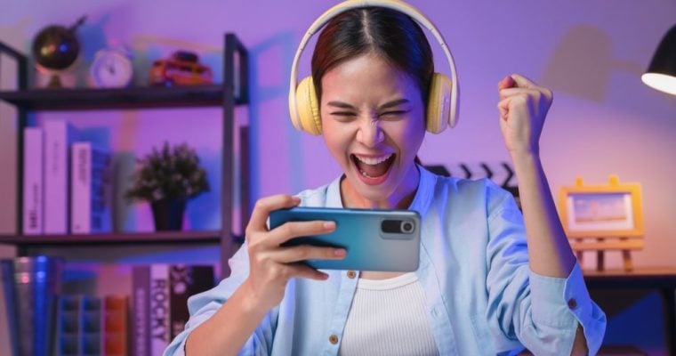 Excited Young Woman Holds iPhone Wearing Yellow Headphones Sitting at Desk Inside Room