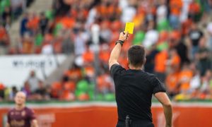 Fans Sit in Soccer Field Stadium While Referee Holds Up A Yellow Card to Player