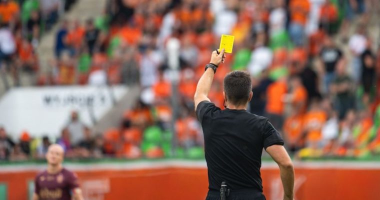Fans Sit in Soccer Field Stadium While Referee Holds Up A Yellow Card to Player