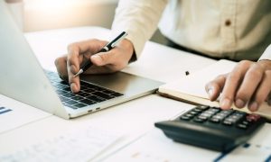 Man in White Shirt Crunches Numbers on Calculator Sitting on Desk with Documents