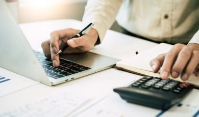 Man in White Shirt Crunches Numbers on Calculator Sitting on Desk with Documents