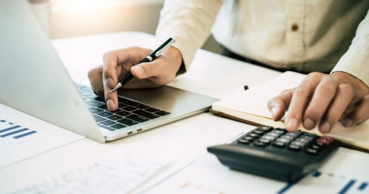 Man in White Shirt Crunches Numbers on Calculator Sitting on Desk with Documents