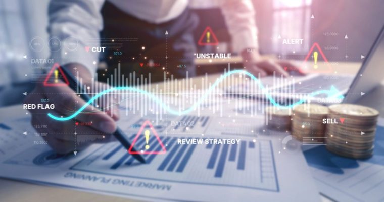 Man Analysis Documents with Bar Graphs In Front of Laptop with Digital Image Overlay of Wave Graph and Warning Icons