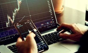 Man Uses Laptop and Cell Phone to Anaylize Financial Graphs While Sitting At Desk