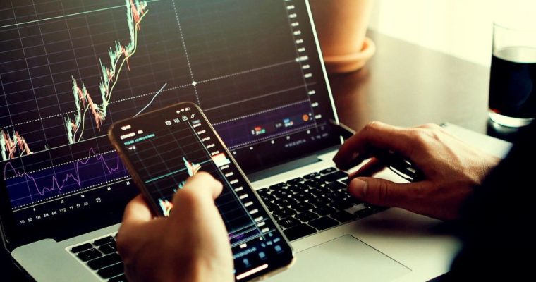 Man Uses Laptop and Cell Phone to Anaylize Financial Graphs While Sitting At Desk