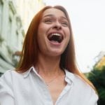 Woman Wearing White Shirt Standing Outdoors Holds Cell Phone With Excited Expression
