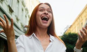 Woman Wearing White Shirt Standing Outdoors Holds Cell Phone With Excited Expression