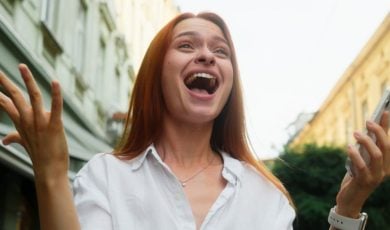 Woman Wearing White Shirt Standing Outdoors Holds Cell Phone With Excited Expression