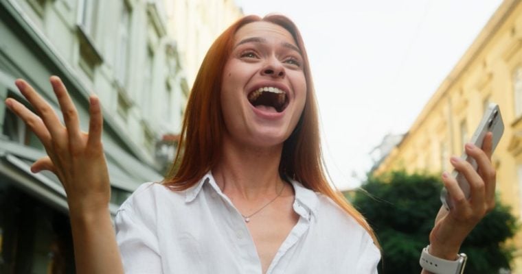 Woman Wearing White Shirt Standing Outdoors Holds Cell Phone With Excited Expression