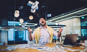 Happy Man Sits In Office In Front of Laptop Throwing Cash In the Air