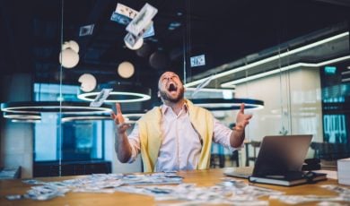 Happy Man Sits In Office In Front of Laptop Throwing Cash In the Air