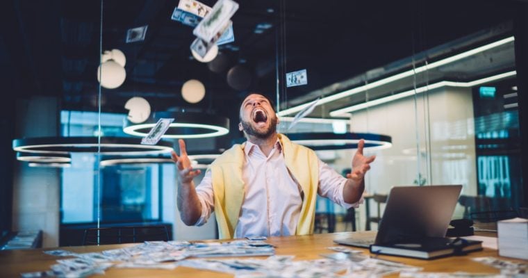 Happy Man Sits In Office In Front of Laptop Throwing Cash In the Air
