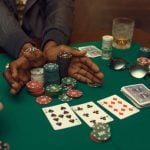 Three Poker Players Sit At Green Game Table with Chips, Playing Cards and Cash on It
