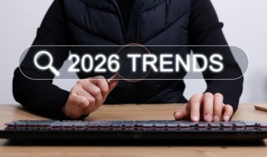 Man Wearing All Black Holds Magnifying Glass Sitting At Desk In Front of Keyboard