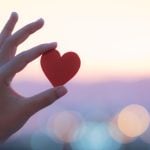 Sunset Colors Blurred Background to Person Holding Up Small Red Heart with One Hand